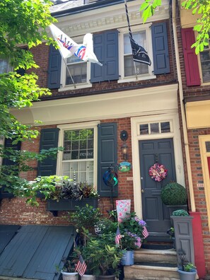 Ein zweistöckiges Backsteingebäude mit einer dunkelblauen Haustür, einer kleinen Veranda mit Blumentöpfen und amerikanischen Flaggen.