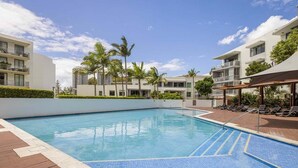 Una piscina rodeada de edificios modernos con balcones, palmeras y un cielo despejado.