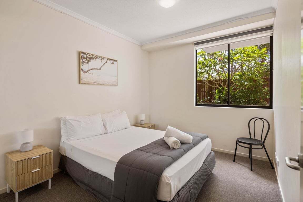 A bedroom with a bed, a nightstand, a chair, a window with a view of trees, and a wall-mounted picture.