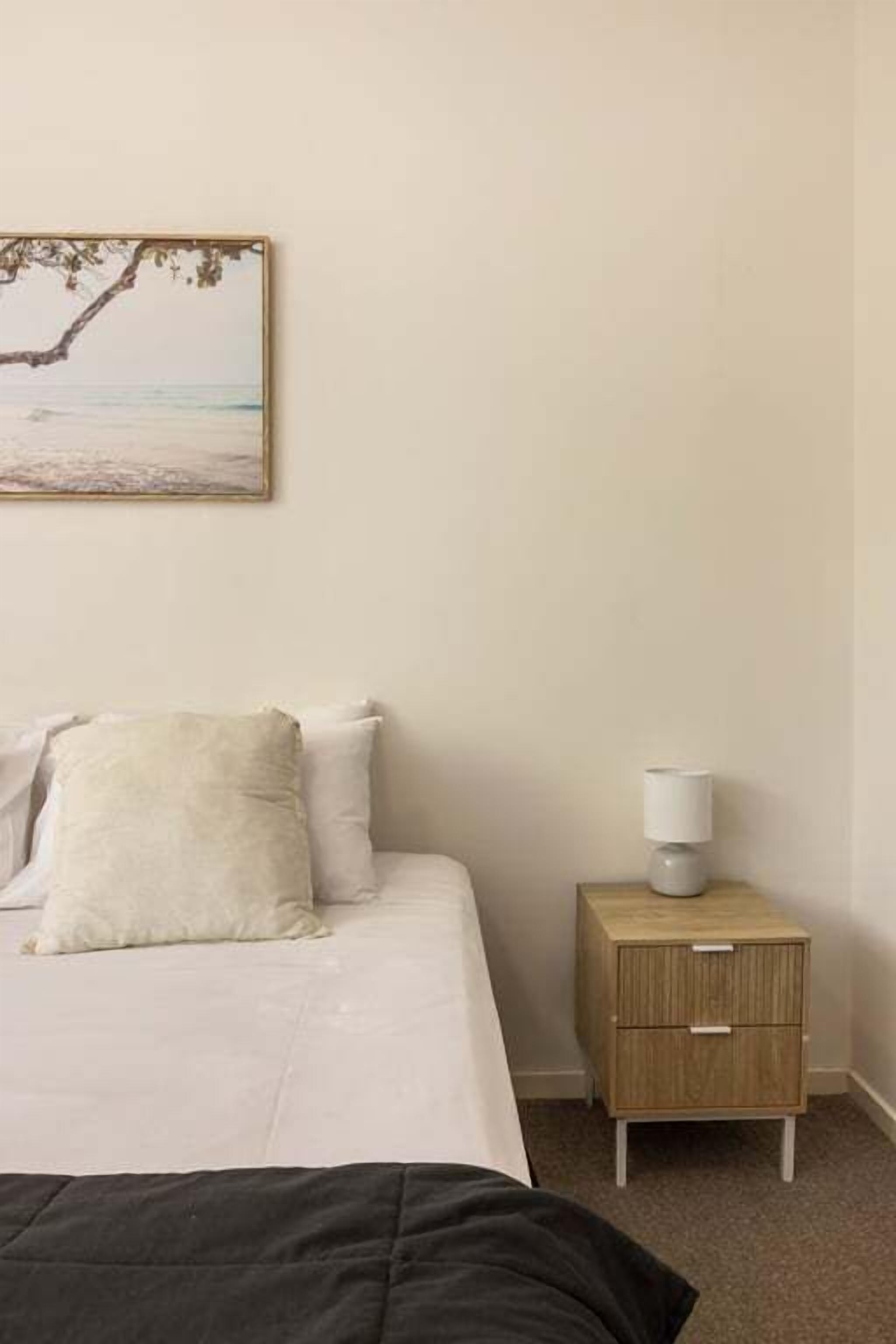 A bedroom with a bed, a nightstand with a lamp, and a wall-mounted picture of a beach scene.
