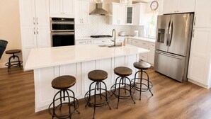 Una cocina moderna con gabinetes blancos, electrodomésticos de acero inoxidable, una isla grande con taburetes e iluminación colgante.