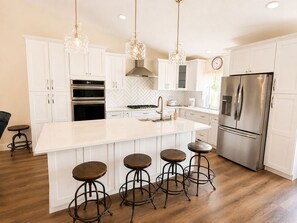 A modern kitchen with white cabinets, stainless steel appliances, a large island with stools, and pendant lighting. - Amazing 4BR Near Shops, Lake Lowell & Wineries (Nampa)
