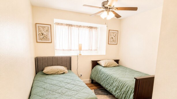 Una habitación pequeña y luminosa con dos camas individuales, un ventilador de techo, una ventana con cortinas traslúcidas y dos cuadros con motivos florales en las paredes.