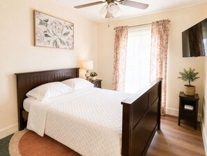 Een slaapkamer met een bed, een plafondventilator, een nachtkastje met een lamp, een potplant en een schilderij aan de muur.