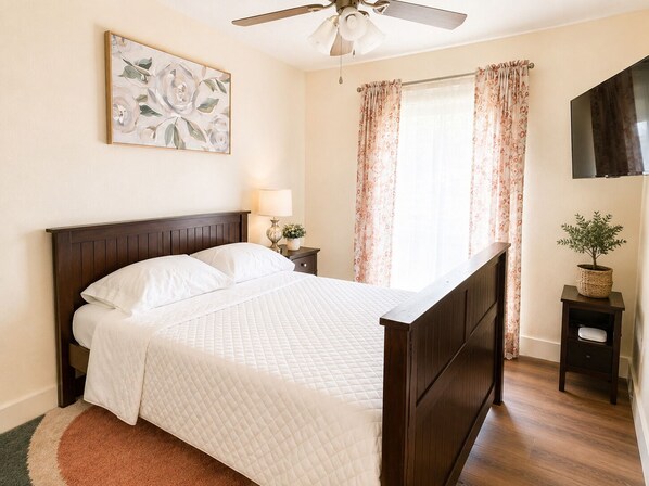 Une chambre à coucher avec un lit, un ventilateur de plafond, une table de chevet avec une lampe, une plante en pot et un tableau accroché au mur.
