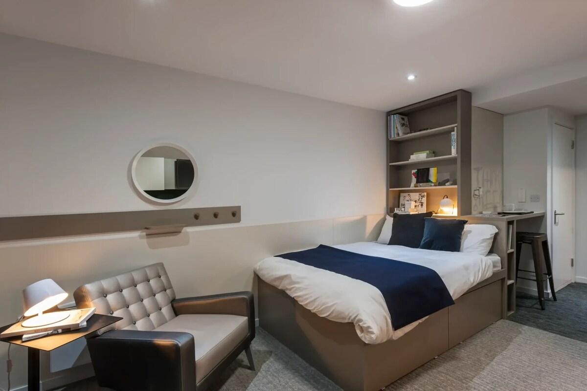 A hotel room with a bed, a desk with a chair, a bookshelf, and a round mirror on the wall.