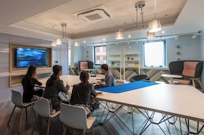 A modern meeting room with a large table, chairs, a TV screen, and shelves with items. - Student Only West Village (Glasgow)