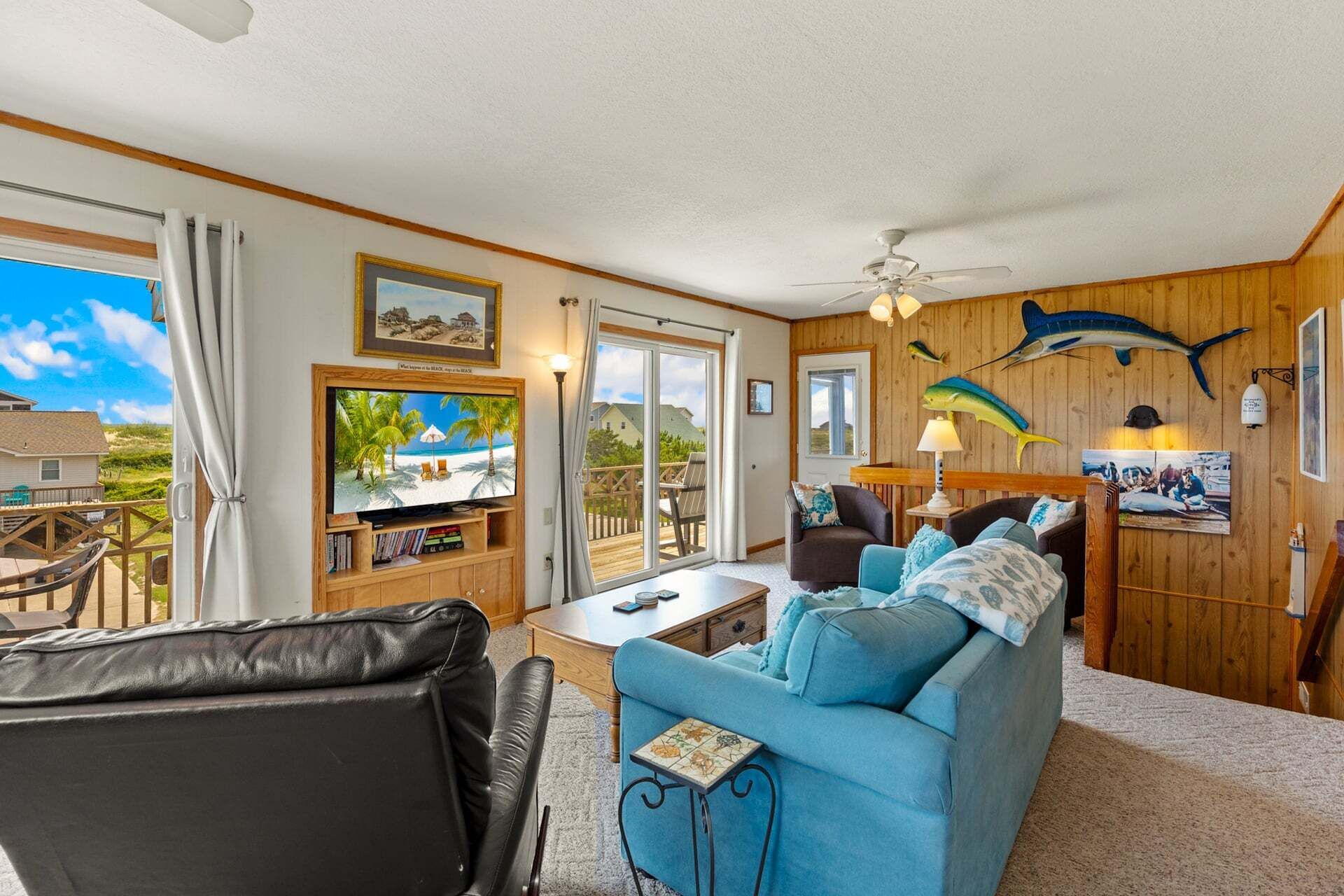 A room with a large TV, a sofa, a chair, a table, and a ceiling fan.