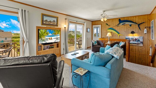 A room with a large TV, a sofa, a chair, a table, and a ceiling fan. - Surfside Rendezvous - Semi-oceanfront  with ocean views, easy beach access, and deck (Nags Head)
