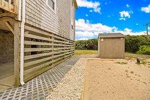 A wooden fence, a shed, and a gravel path leading to a house with a window. - Surfside Rendezvous - Semi-oceanfront  with ocean views, easy beach access, and deck (Nags Head)