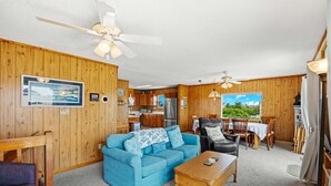 A cozy cabin interior with a blue sofa, wooden coffee table, and a dining area with a view. - Surfside Rendezvous - Semi-oceanfront  with ocean views, easy beach access, and deck (Nags Head)