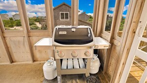 A portable Weber grill with two propane tanks, set up in a wooden structure with a view of houses in the background. - Surfside Rendezvous - Semi-oceanfront  with ocean views, easy beach access, and deck (Nags Head)