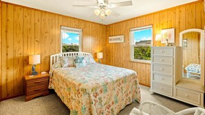 A bedroom with wooden walls, a bed with floral bedding, a nightstand, a dresser, and a ceiling fan. - Surfside Rendezvous - Semi-oceanfront  with ocean views, easy beach access, and deck (Nags Head)
