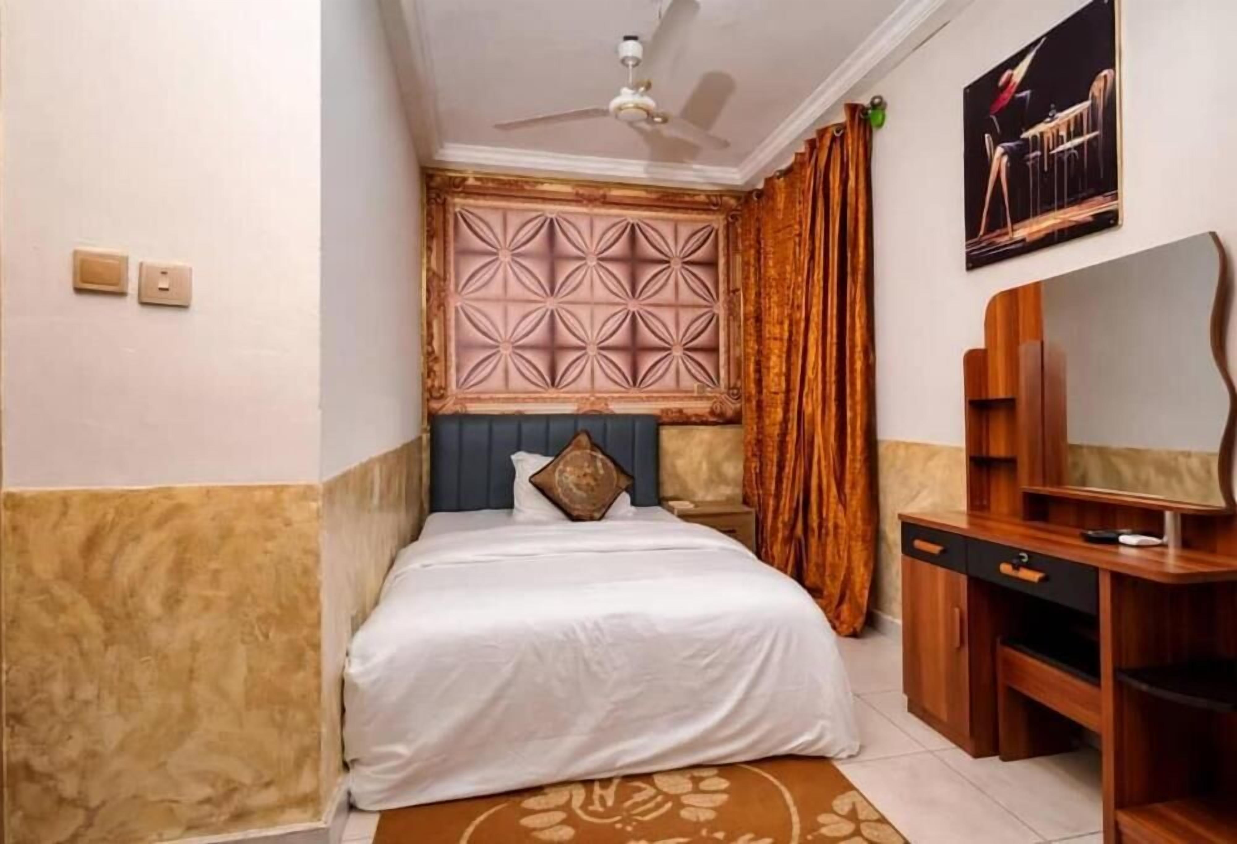 A bedroom with a bed, a desk, a ceiling fan, a wall-mounted TV, and a decorative headboard.