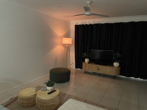 Ein Wohnzimmer mit Deckenventilator, einem Flachbildfernseher auf einer Holzeinrichtung, einer Stehlampe, zwei runden Hocker und einem kleinen Topfpflanzen.