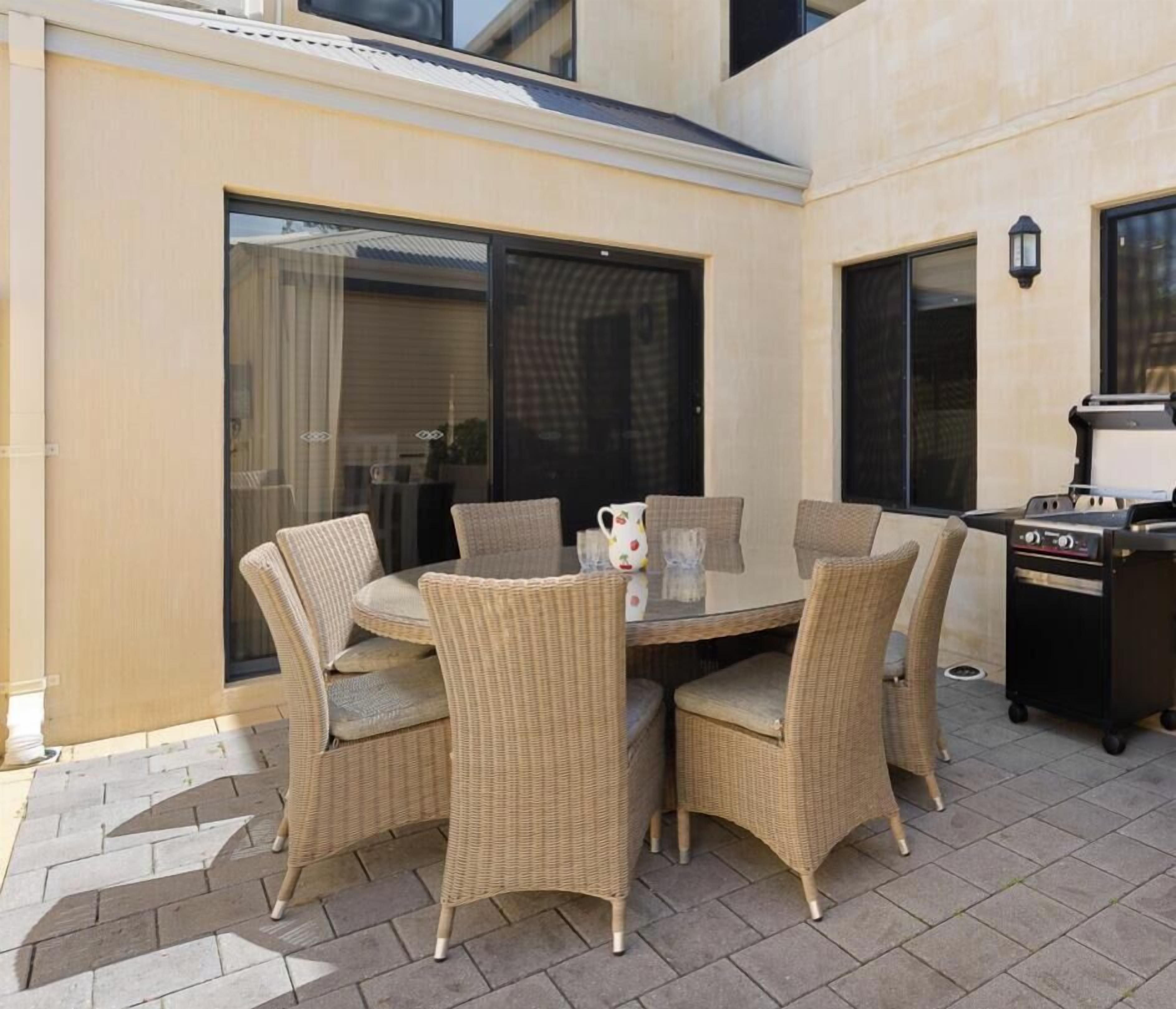 Une terrasse avec une table en verre et des chaises en osier, un barbecue et une grande fenêtre.