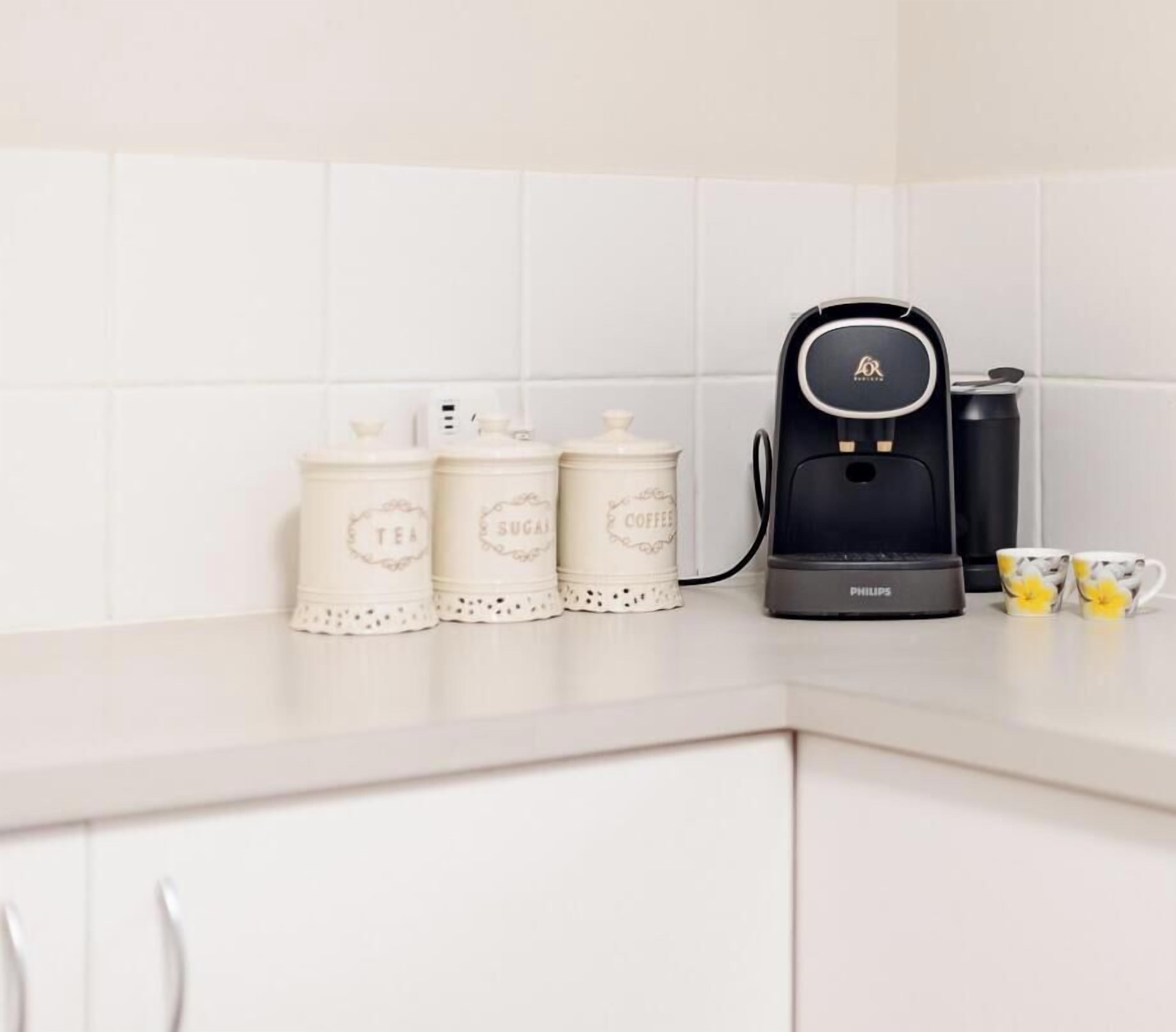 Un comptoir de cuisine avec une machine à café, deux récipients étiquetés « THÉ », « SUCRE » et « CAFÉ », et une tasse.