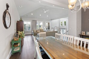 Ein geräumiger Essbereich mit einem langen Holztisch und weißen Geländern, ein grüner Beistelltisch mit Korb und Blick auf ein Wohnzimmer mit Sofa und Fenster.
