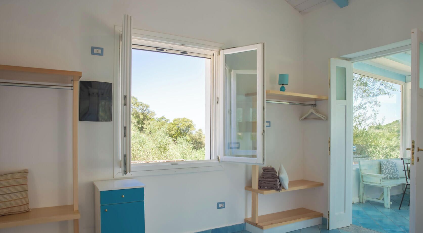 A room with a window, a desk, a chair, and a small shelf with towels.
