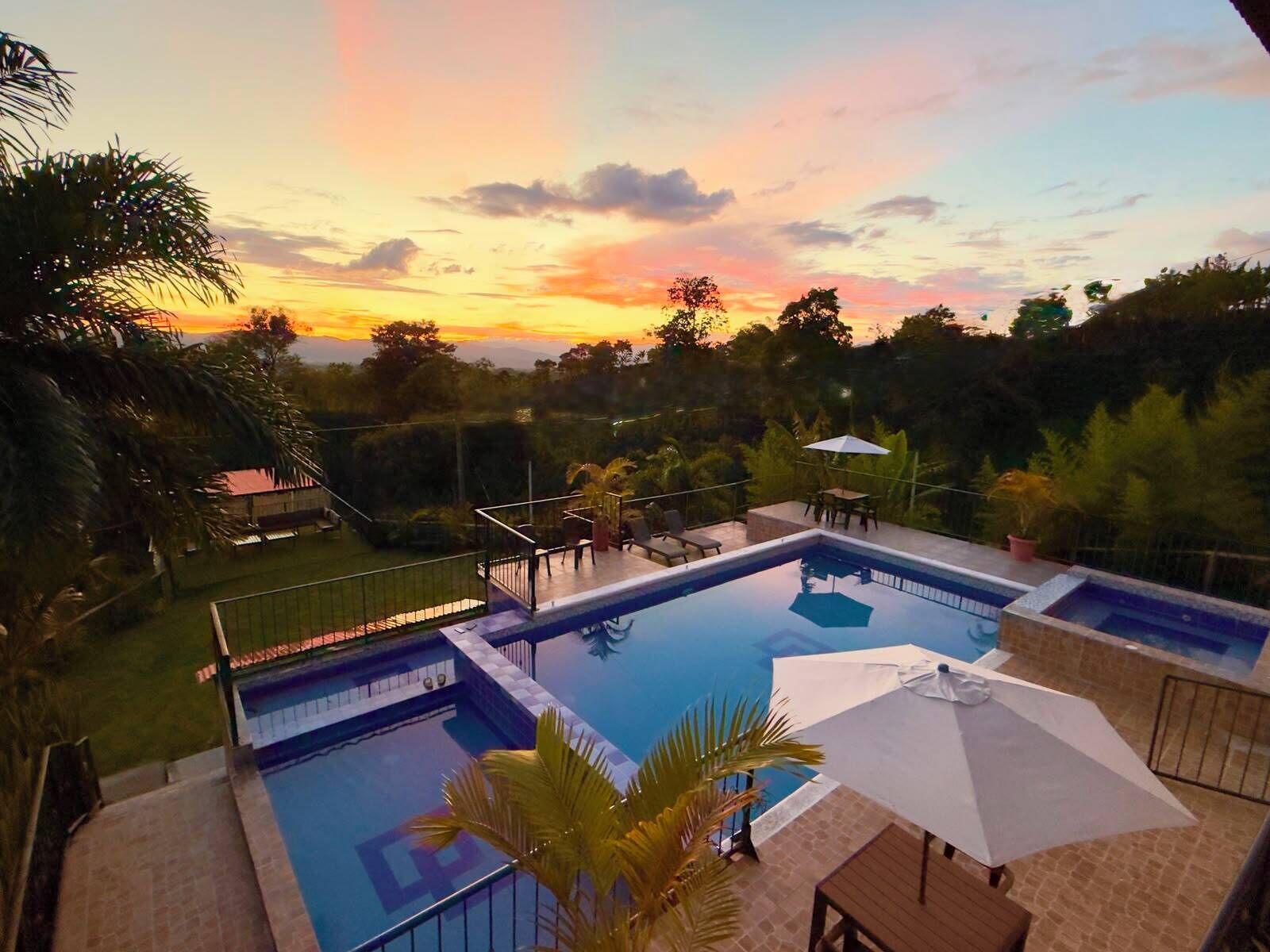 A swimming pool with a view of the sunset, surrounded by greenery and a patio area with seating.