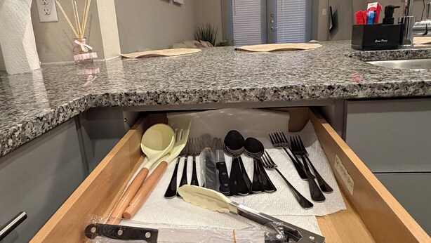 Una cocina con encimera de granito, un organizador de cajones con utensilios, un ventilador de techo y una ventana con persianas.