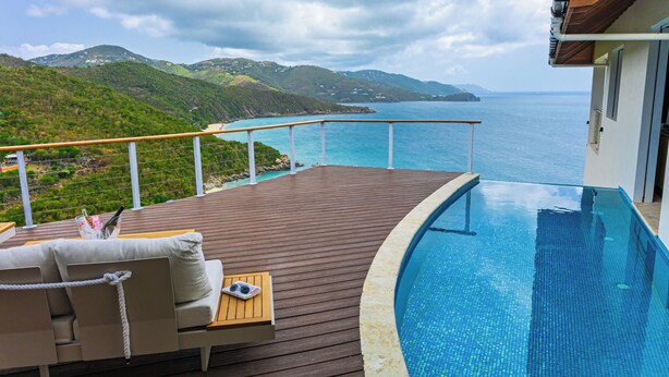 Ein Poolbereich mit Holzterrasse, Liegestuhl und Blick auf das Meer und die Hügel.