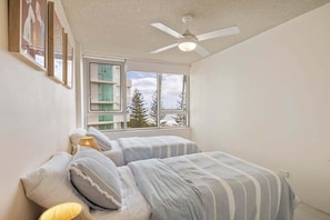 Ein Schlafzimmer mit zwei Betten, einem Deckenventilator, einem Fenster mit Blick auf Bäume und gerahmten Bildern an der Wand.