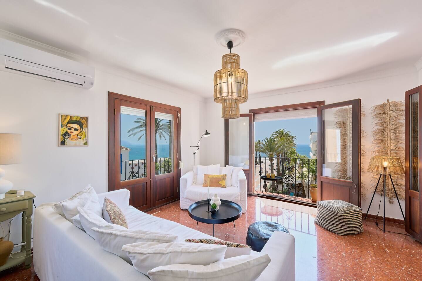 A hotel room with a white sofa, a round coffee table, a lamp, a painting on the wall, and a view of the sea through large glass doors.