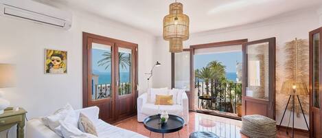 A hotel room with a white sofa, a round coffee table, a lamp, a painting on the wall, and a view of the sea through large glass doors.
