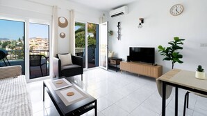 Una sala moderna con televisor, sofá y comedor, ventanas amplias y un balcón con vistas.