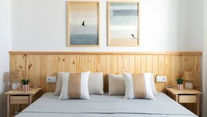 Un dormitorio con cabecera de madera, dos cuadros enmarcados que muestran un velero y un pájaro, una cama con ropa de cama a rayas, y mesitas de noche con lámparas y plantas.