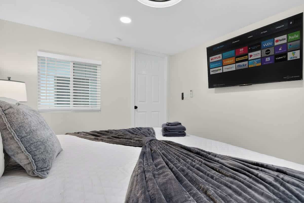 A hotel room with a bed, a TV mounted on the wall displaying various streaming services, a window with blinds, and a door.