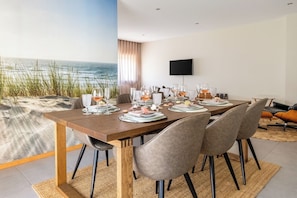 Ein Esszimmer mit Blick auf den Strand, einem Flachbildfernseher an der Wand und einem für sechs Personen gedeckten Tisch mit Gläsern und Tellern.
