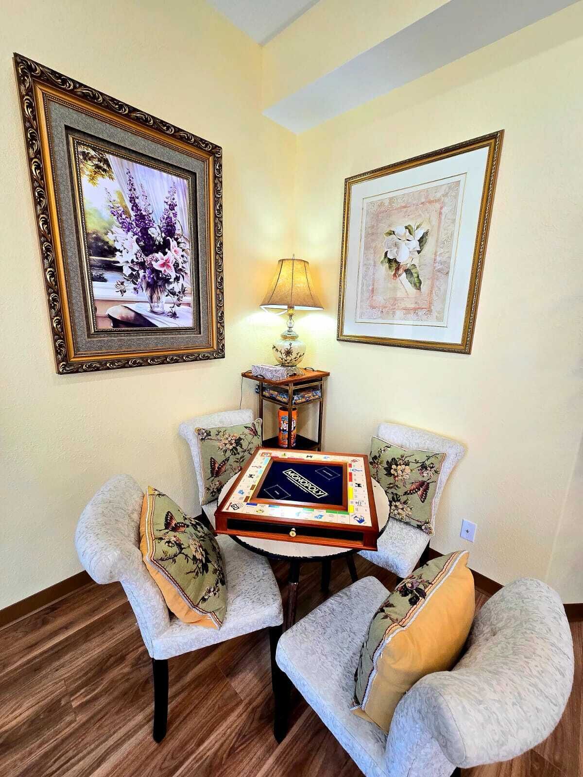 A room with a Monopoly board game on a table, two chairs with floral cushions, a lamp, and two framed pictures on the wall.