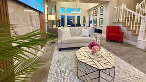 A modern living room with a sofa, a coffee table, and a red chair. There is a staircase with white railings and a painting on the wall. - Relaxing & Luxurious Private Oasis w heated Pool/Spa 10 min from Las Vegas Strip (Henderson)