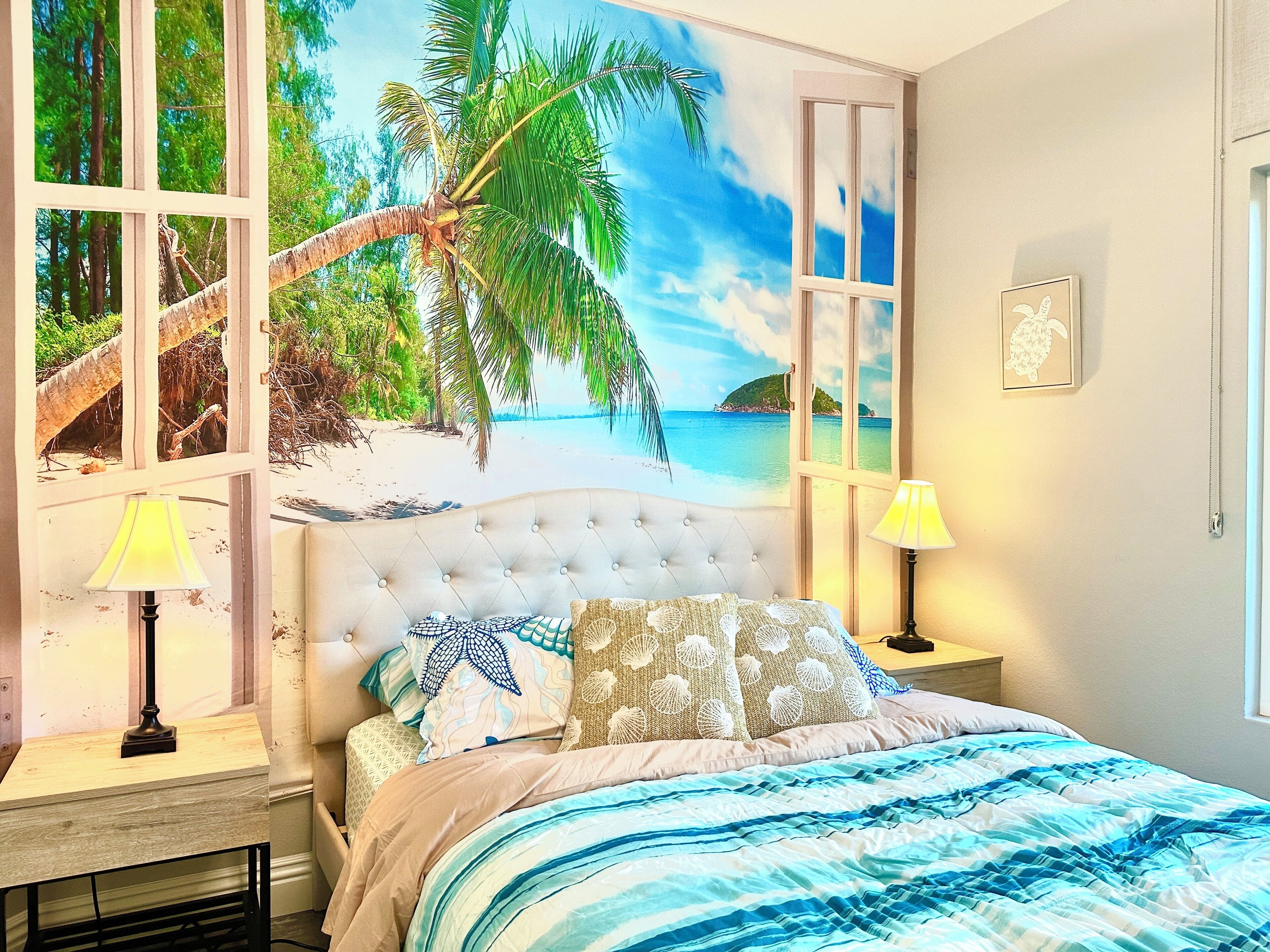 Un dormitorio con un mural de playa, un cabecero capitonado, una cama con ropa de cama estampada, dos lámparas de noche y un cuadro en la pared.
