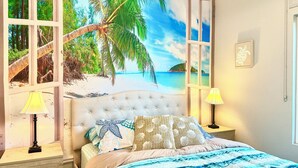 A bedroom with a beach mural, a tufted headboard, a bed with patterned bedding, two bedside lamps, and a framed picture on the wall. - Relaxing & Luxurious Private Oasis w heated Pool/Spa 10 min from Las Vegas Strip (Henderson)