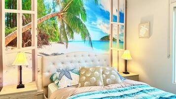 Un dormitorio con un mural de playa, un cabecero capitonado, una cama con ropa de cama estampada, dos lámparas de noche y un cuadro en la pared.