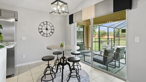Eine moderne Küche mit einem runden Esstisch, Barhocker, einer großen Wanduhr und Blick durch eine Glastür auf eine Außenpatio.