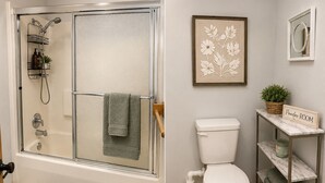 Un baño con ducha de vidrio, inodoro, una obra de arte floral enmarco, una planta pequeña en maceta y un estante con un letrero que dice 'Powder-Room'.