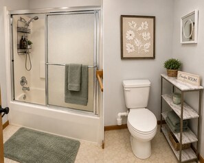 A bathroom with a glass-enclosed shower, a toilet, a framed floral artwork, a small potted plant, and a shelf with a sign reading 'Powder-Room'. - Cozy 3-bedroom house in charming Cedar Falls (Cedar Falls)