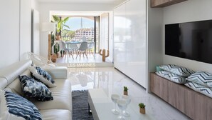A modern living room with a sofa, coffee table, and TV. There is a balcony with a view of buildings and a pool. - Escapade des Marines - Welkeys (Cogolin)