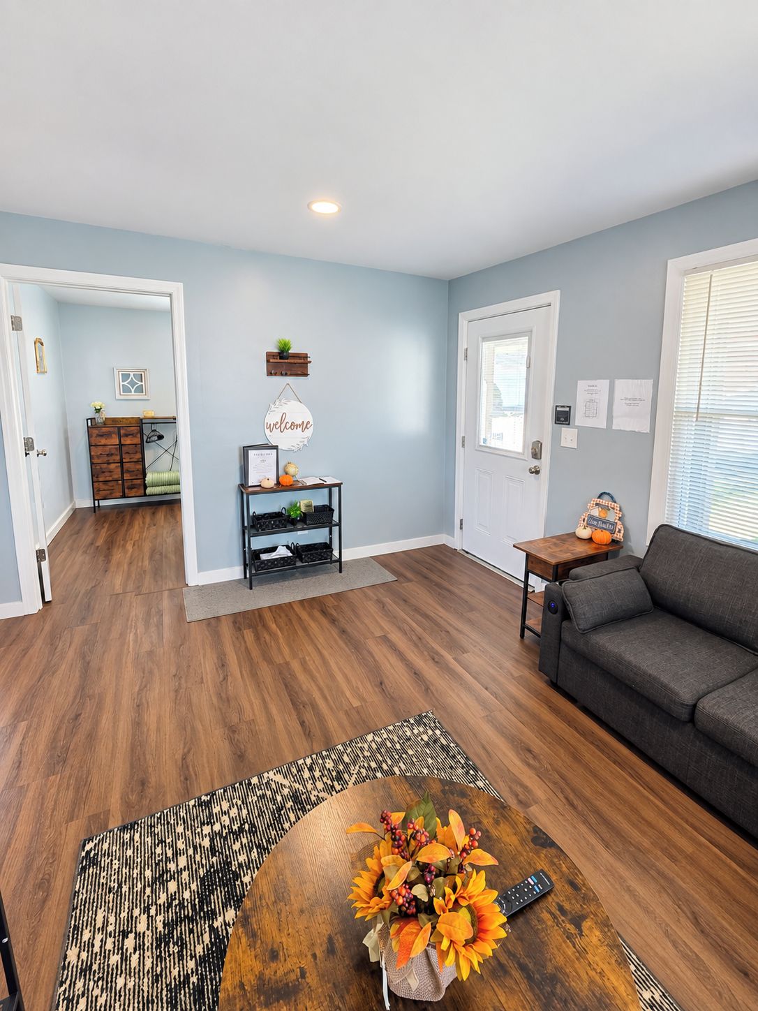 A living room with a sofa, a coffee table with a floral arrangement, a wooden floor, and a door leading to another room.