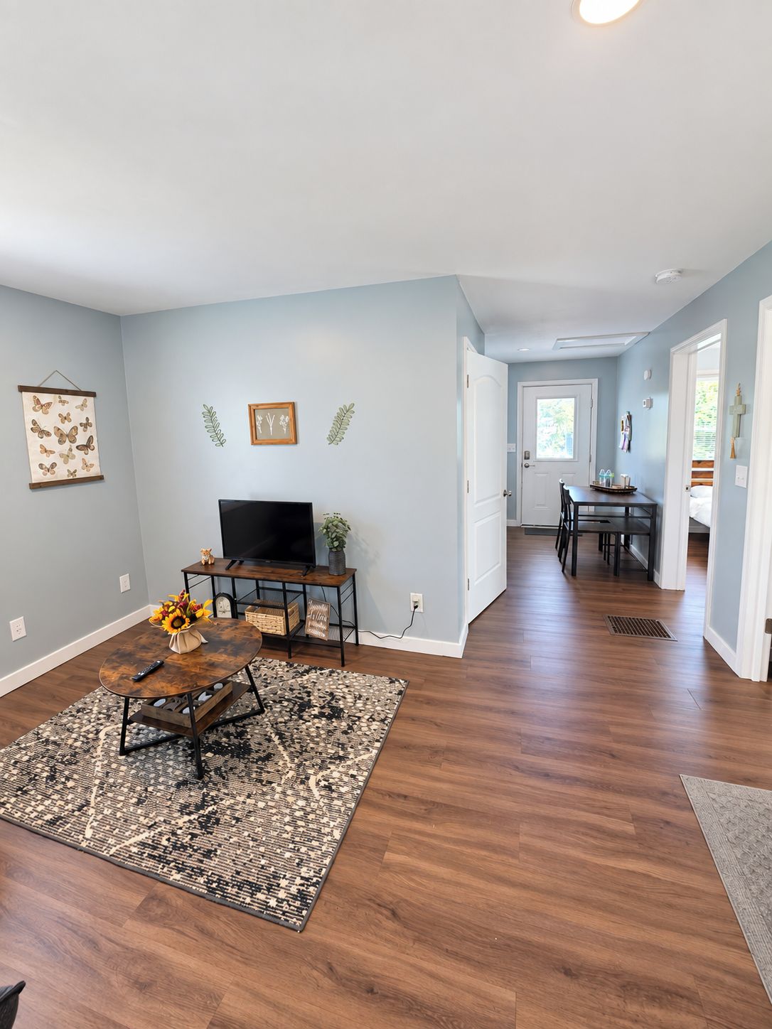 A spacious living area with hardwood flooring, a flat-screen TV, a coffee table with a vase of flowers, and a dining area with a table and chairs.