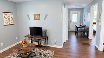 A spacious living area with hardwood flooring, a flat-screen TV, a coffee table with a vase of flowers, and a dining area with a table and chairs.