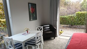 Une pièce avec une table blanche, une chaise noire, un lit recouvert d’une couverture rouge, un tableau encadré accroché au mur et une vue sur le jardin par la fenêtre.