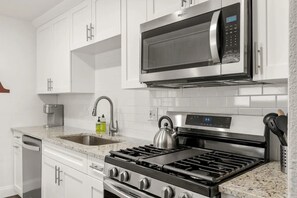 Una cocina con armarios blancos, un microondas de acero inoxidable, una estufa a gas y un hervidor.