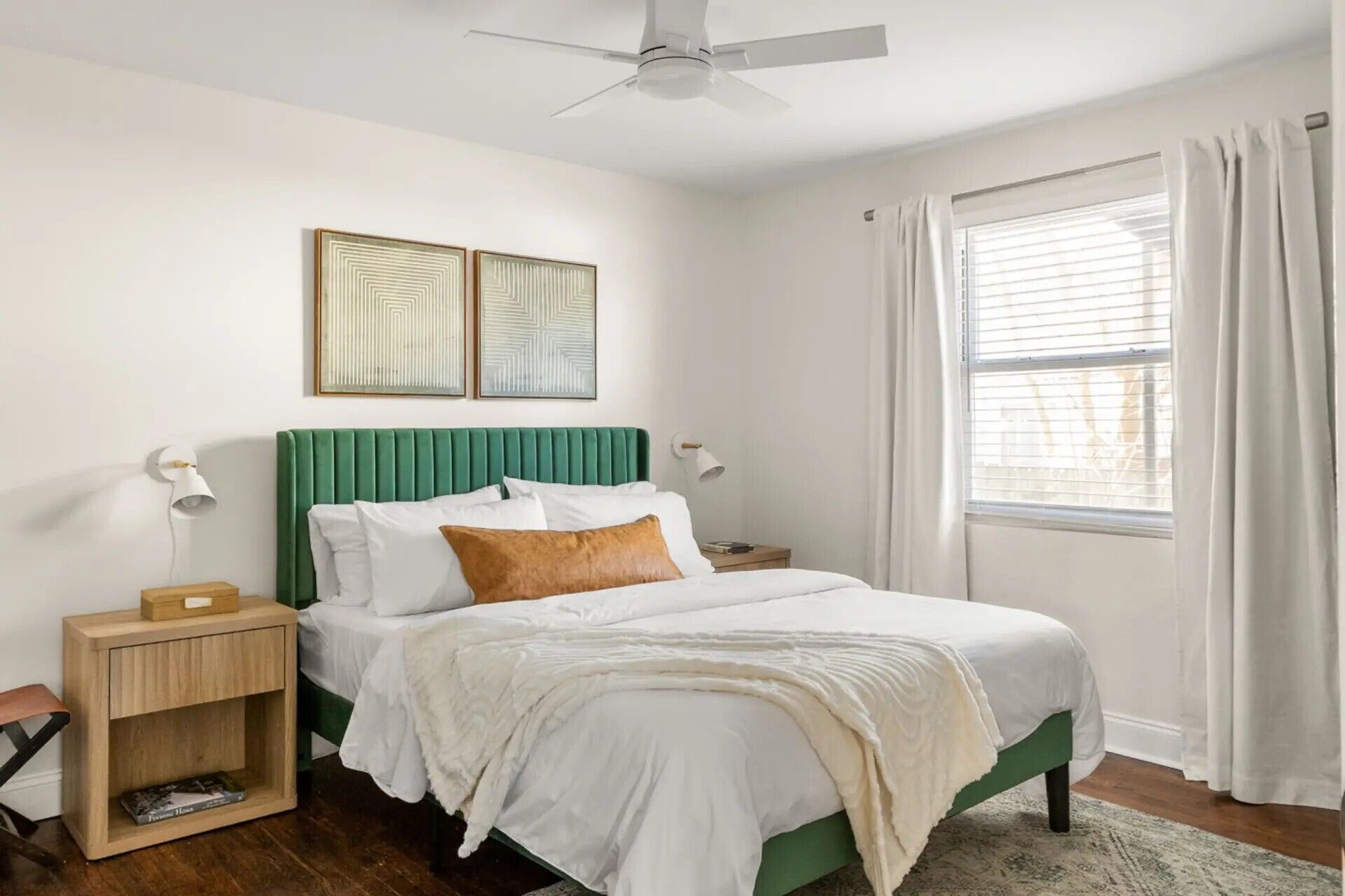 A bedroom with a bed, a nightstand, a ceiling fan, two wall-mounted lamps, and a window with curtains.