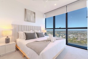 Ein modernes Schlafzimmer mit einem großen Bett, einem Nachttisch, einer Lampe und Blick auf die Stadt durch große Fenster.