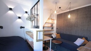 Un espace de vie moderne et compact comprenant un lit, un canapé, une petite table et une étagère intégrée où sont rangés des livres et des objets décoratifs.